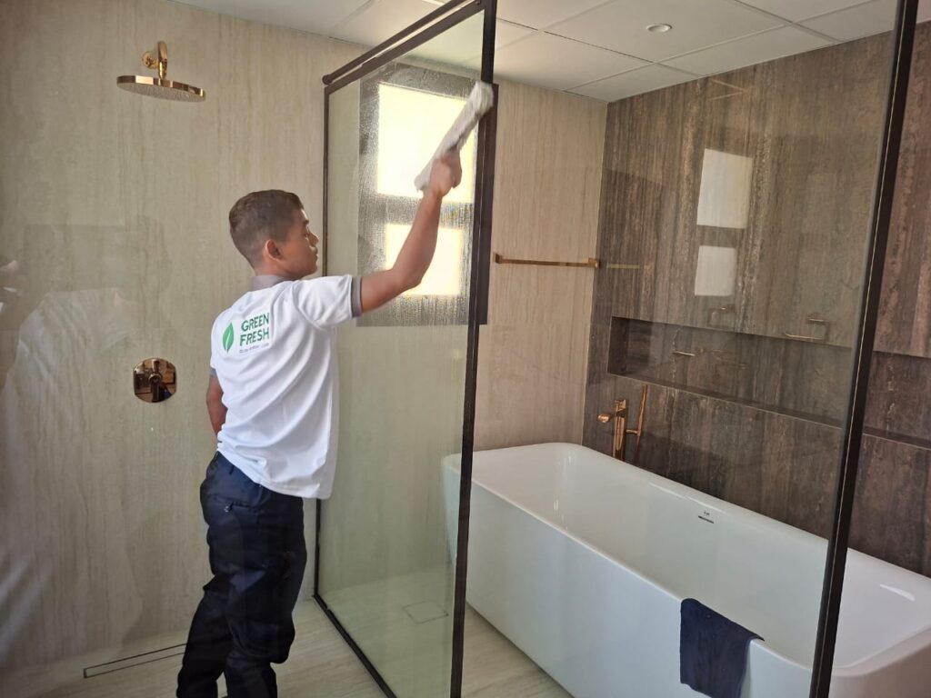 Cleaner wearing a "Green Fresh" uniform using a squeegee to clean the glass shower enclosure in a luxurious bathroom with a freestanding tub.