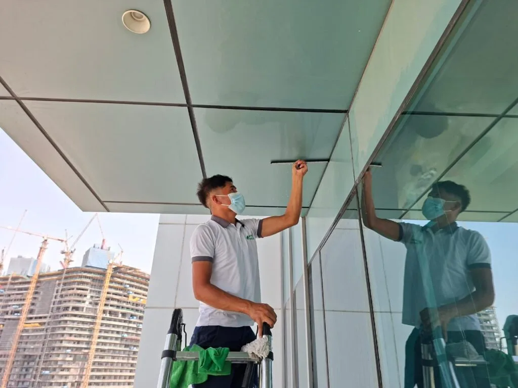 Professional cleaner on a ladder using a squeegee to clean a large exterior glass panel on a modern high-rise building.