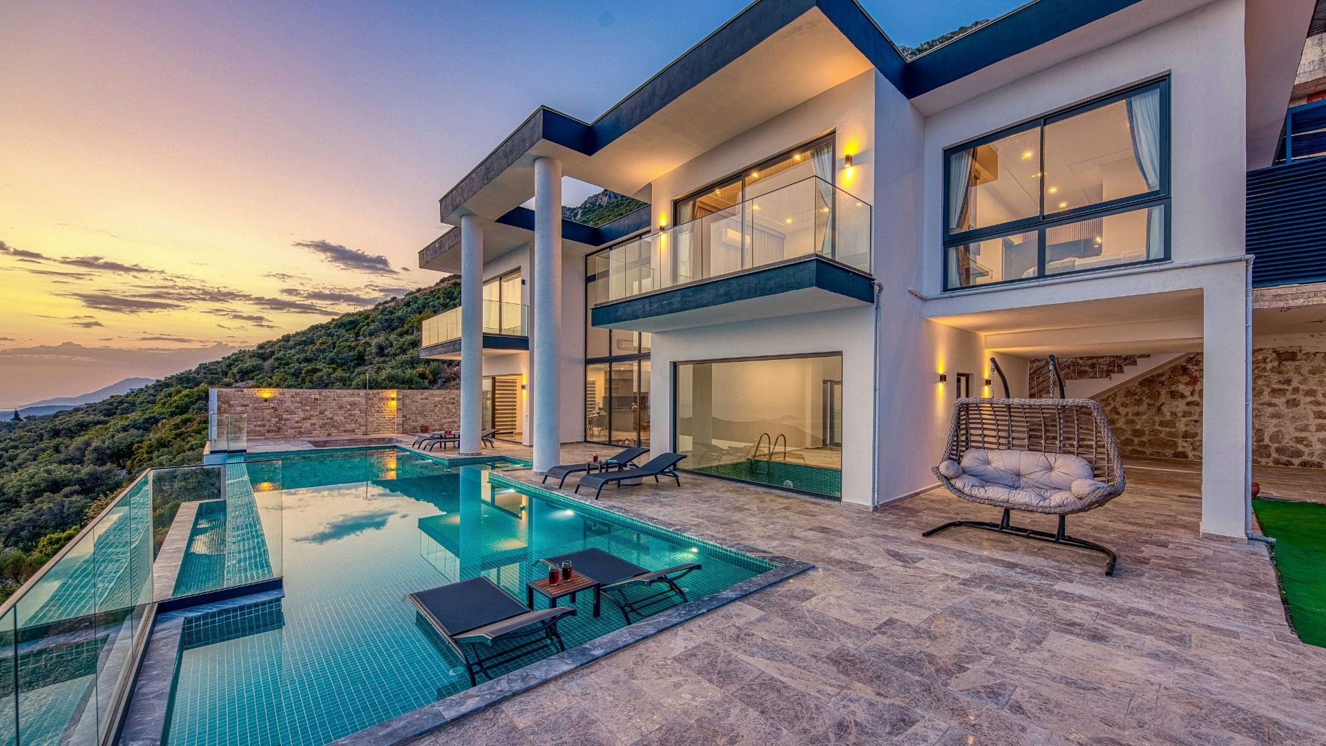 Luxury hillside villa with an infinity pool, glass balconies, outdoor lounge chairs, and panoramic mountain views at sunset.