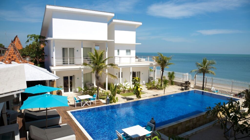 Modern beachfront villa with a large blue swimming pool, palm trees, outdoor seating, and ocean views on a sunny day.