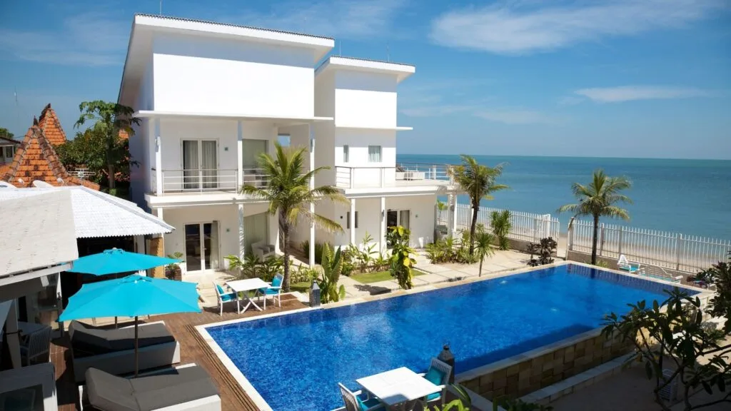 Villa cleaning services 4 Modern beachfront villa with a large blue swimming pool, palm trees, outdoor seating, and ocean views on a sunny day.